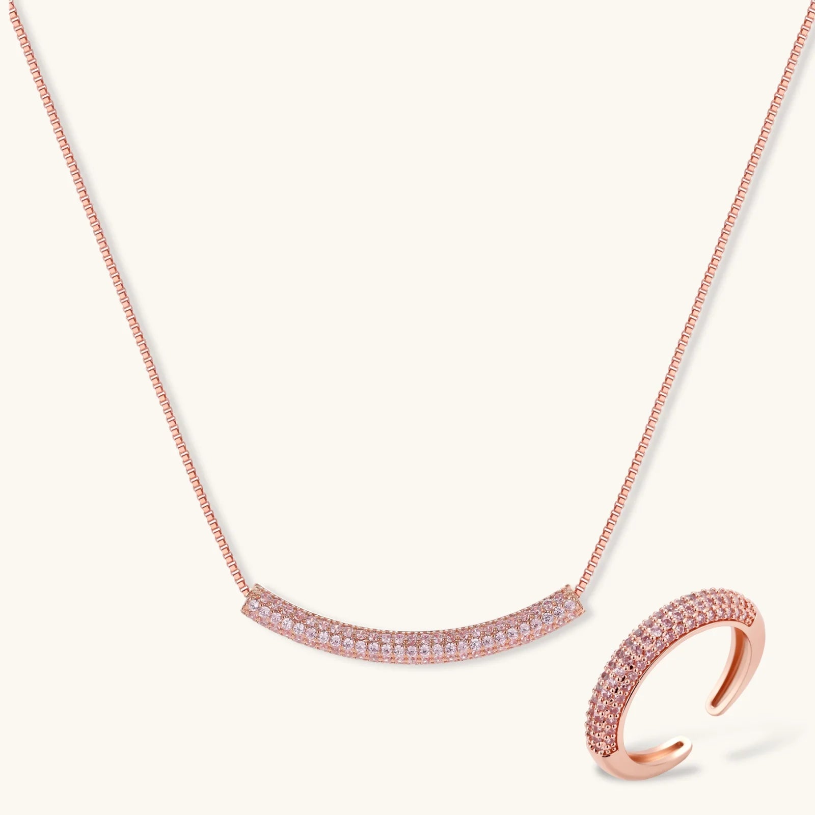 Rose gold jewelry set with pavé cubic zirconia stones including curved necklace, hoop earrings, and adjustable ring on cream background.