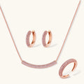 Rose gold jewelry set with pavé cubic zirconia stones including curved necklace, hoop earrings, and adjustable ring on cream background.