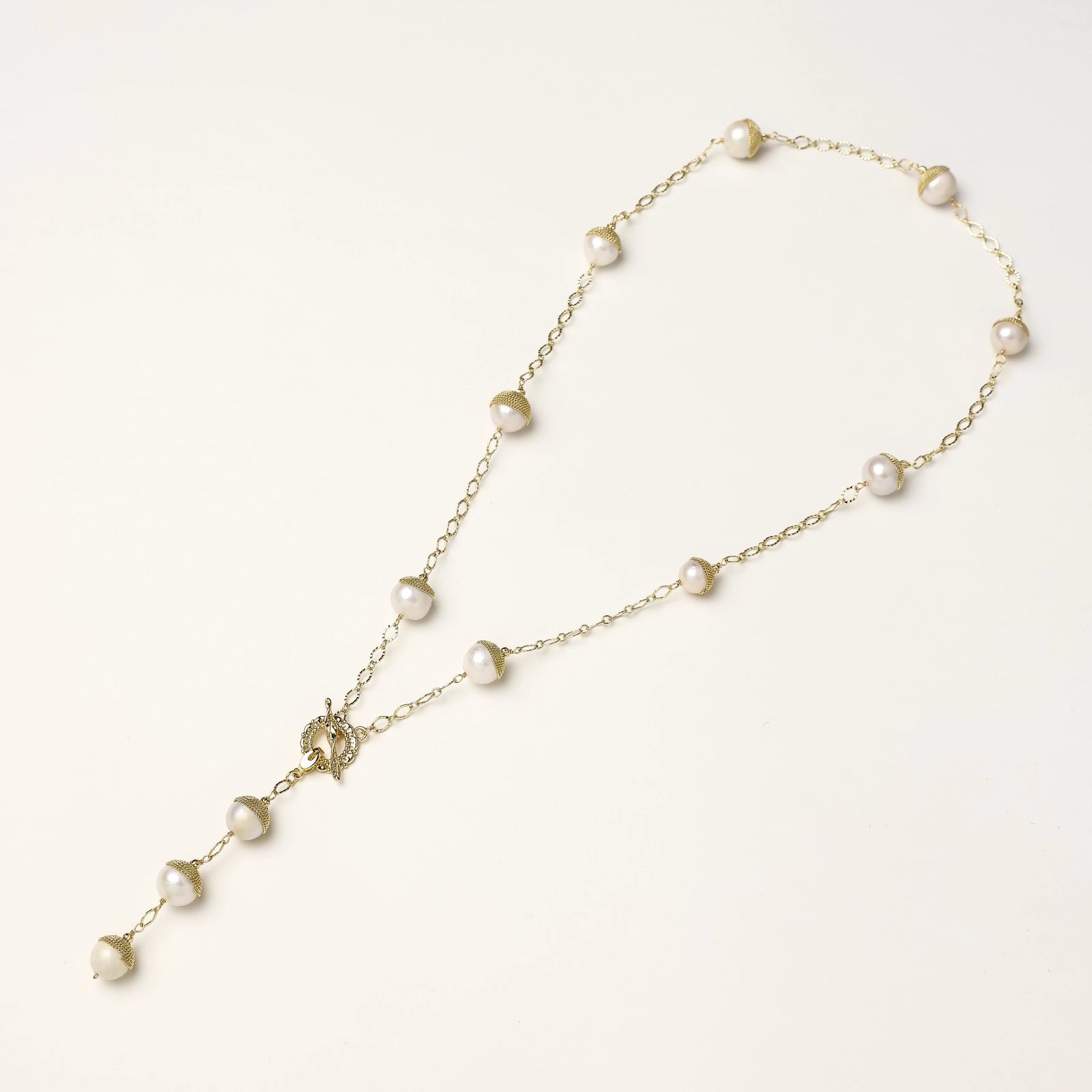 Gold-plated lariat necklace with baroque pearls and textured caps on cream background, showing adjustable Y-shaped design.